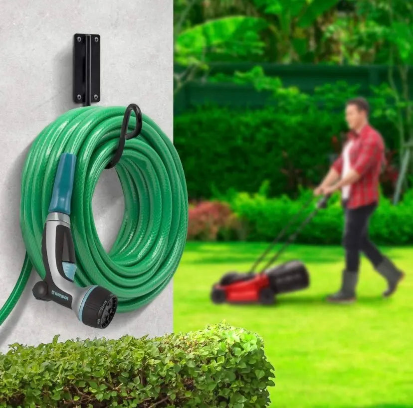 Green garden hose coiled on a wall-mounted holder with a person mowing a lawn in the background.