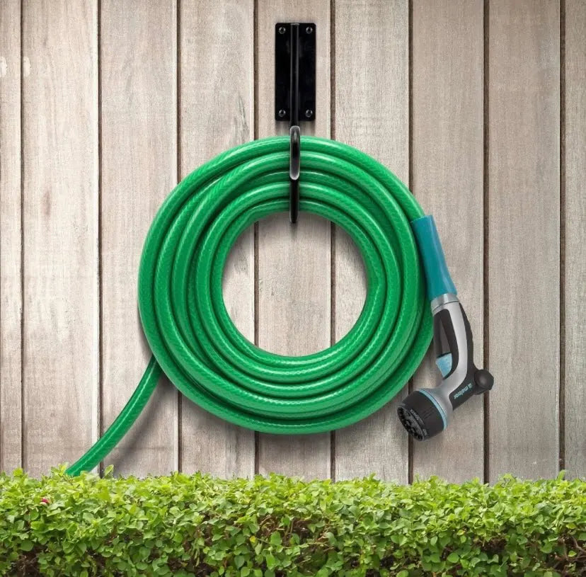 Green garden hose coiled on a wall-mounted holder with a spray nozzle attached, against a wooden fence background.